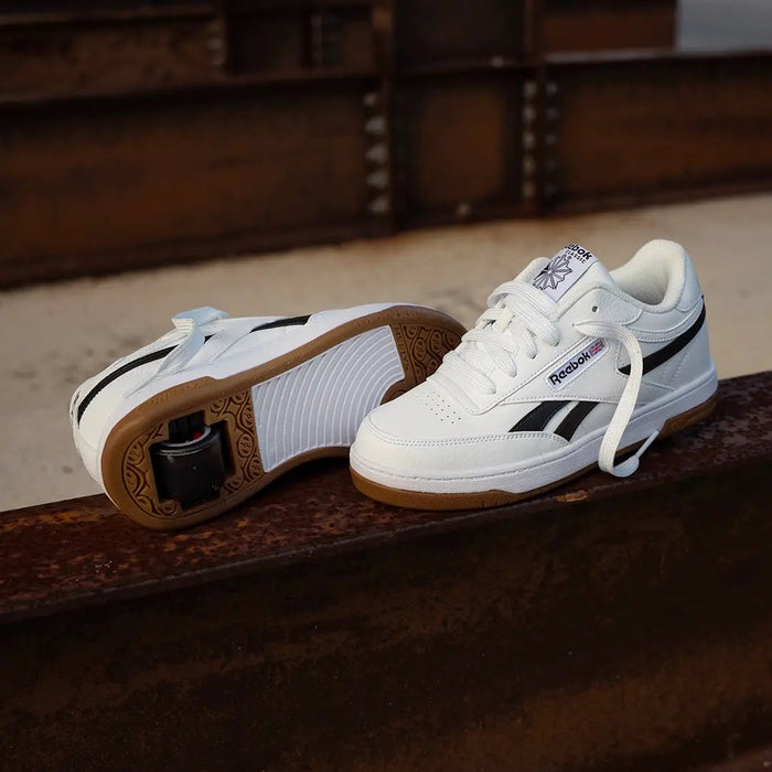 A pair of Reebok low-top sneakers in white with black stripes and brown gum soles, displayed on a rusty metal beam with an industrial background.