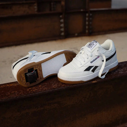 A pair of Reebok low-top sneakers in white with black stripes and brown gum soles, displayed on a rusty metal beam with an industrial background.