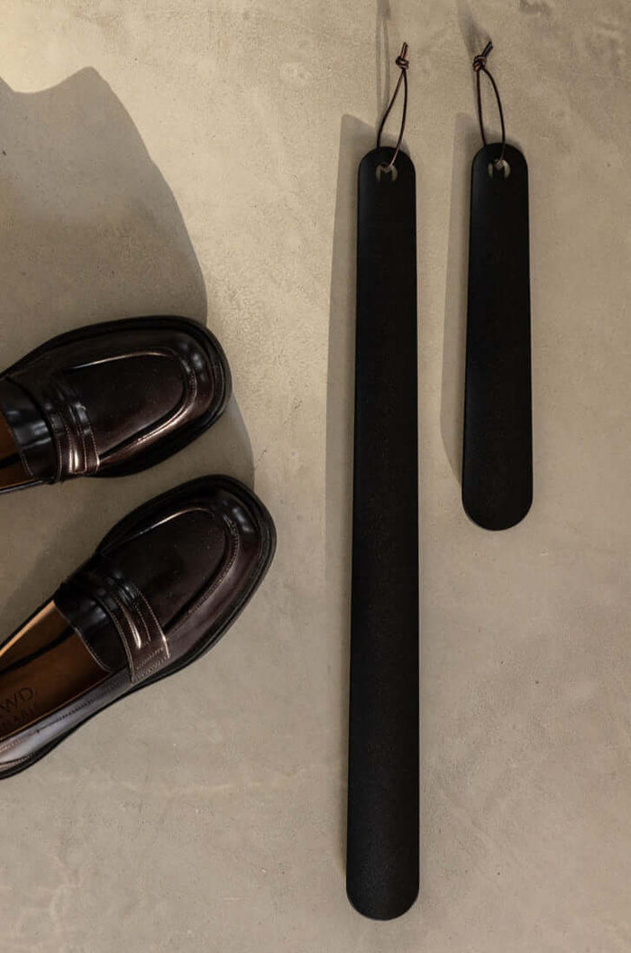 Eleganter Schuhlöffel TILO aus Metall, minimalistische Form neben glänzenden Schläppschuhen auf Betonboden.